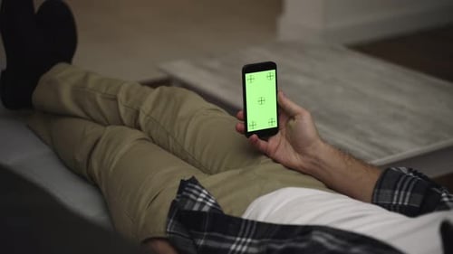 Man Resting on a Couch at Home Using Smartphone with Green Mockup Screen