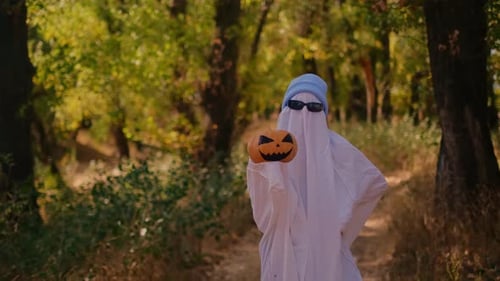 Person in Ghost Costume Holding Jack-O-Lantern in Woods