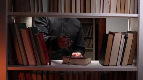 Priest Holding Reading Glasses with Holy Bible and Rosary on Bookshelf