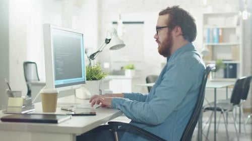 Bearded Creative Man Works on Code While Sitting at His Desk. His Office is Light and Modern.