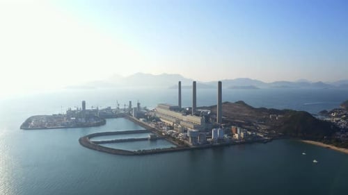 Drone shot, traveling to the left, of a big coastal coal-fired power plant on an island next to a vi