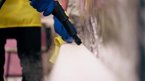 Close Up in an Apartment Cleaner in a Yellow Apron Washes a White Radiator with a Steam Generator