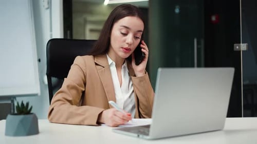 Businesswoman Making Notes While Having Conversation on Smartphone in Office