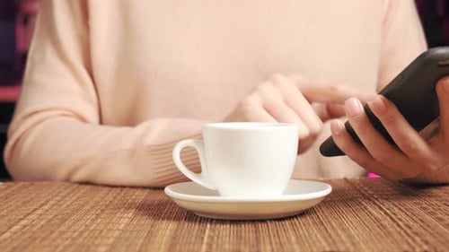 A Girl in a Jacket with a Mobile Phone in Her Hands Drinks Coffee in a Cafe