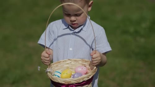 Lustiges kleines Jungenkind hält Osterkorb mit bunten Eiern in Händen und posiert vor der Kamera, sonnig, warm