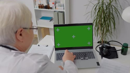 Doctor working in clinic office on laptop with green screen mockup.