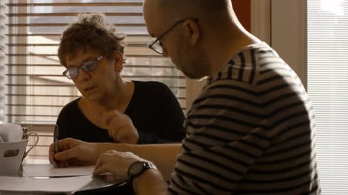 Elderly Woman Signing Documents With Help From Adult Man At Home On Spring Afternoon Medium Shot