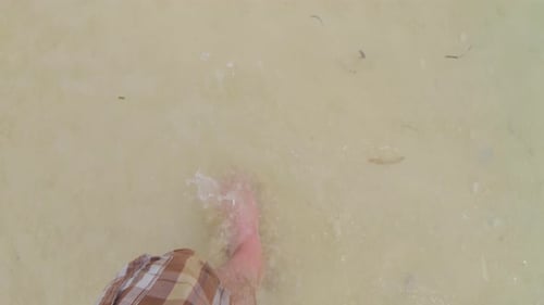 A male's bare feet walking on a sandy beach, with gentle sea waves lapping at his ankles.