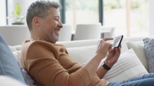 Smiling Mature Man Relaxing on Sofa Using Smartphone at Home