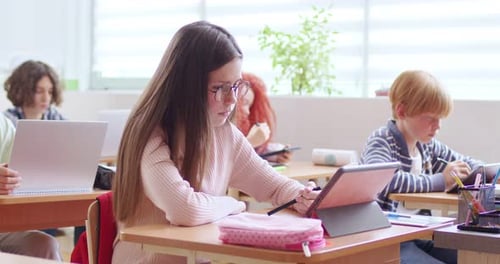 Caucasian Schoolgirl in Glasses Sitting at Desk in Classroom Writing and Using Tablet Device for