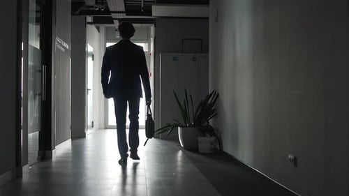 Rear view. The silhouette of a businessman in formal suit walks along a dark corridor of an office