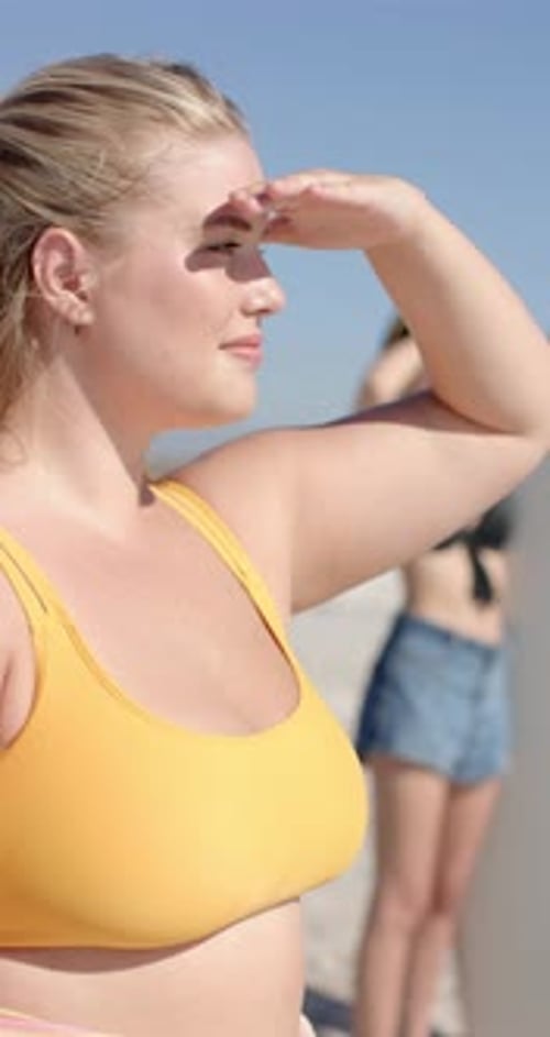 Vertical video: Woman in yellow sports bra looking into distance, enjoying sunny beach day