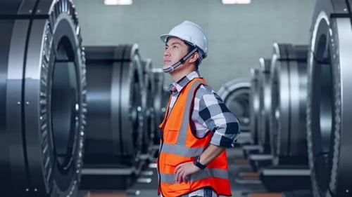 Side View Of Asian Male Engineer Looking Around With Arms Akimbo In Metal Factory