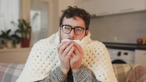 Aria, un homme portant des lunettes, luttant contre les maux saisonniers, tousse et éternue, recouvert d'une couette à la maison