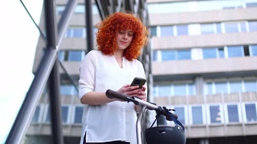 Young Woman with an Electric Scooter Using Her Phone in the City