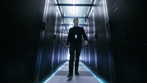 Following Shot of IT Engineer Walking Through Data Center Corridor with Rows of Rack Servers.