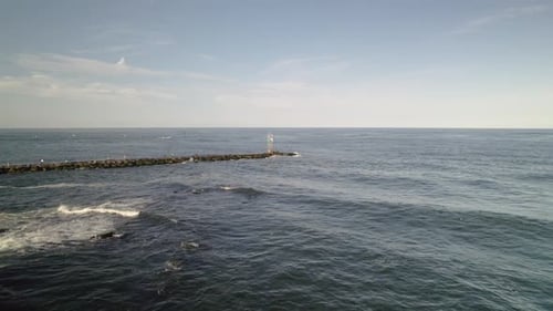 Aerial drone shot pan around light house bouy on rock jetty as man fishes with heavy lens flare