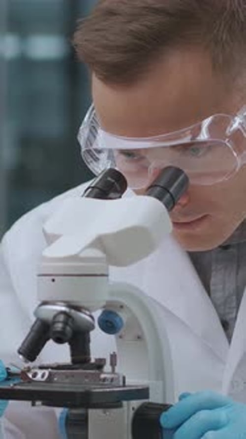 Scientist Wearing Protective Goggles and Gloves Closely Examining Samples Through a Microscope in a