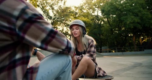 A Girl and a Guy in Plaid Shirts Sit on a Skateboard and Chat in a Skate Park A Blonde Girl in a