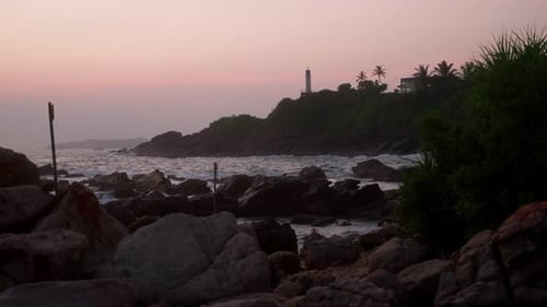 Lighthouse Stands on Green Hill Overlooks Ocean As Waves Crash Against Rocky Shore at Pink Sunset