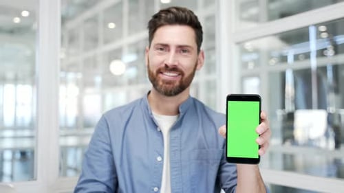 Smiling businessman presenting mobile phone with green screen standing in modern business office.