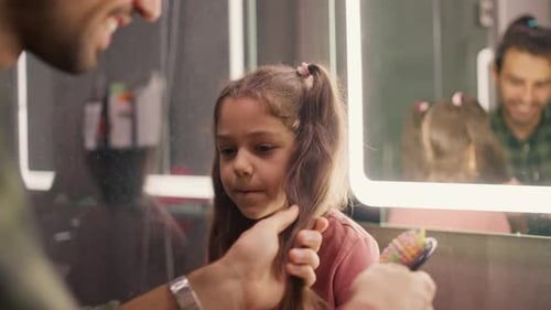 Happy Brunette Man in a Green Checkered Shirt Combs His Little Daughter's Brunette Girl Hairstyle
