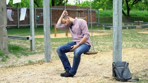 Worried man listens to music on cellphone sitting on swing in city park