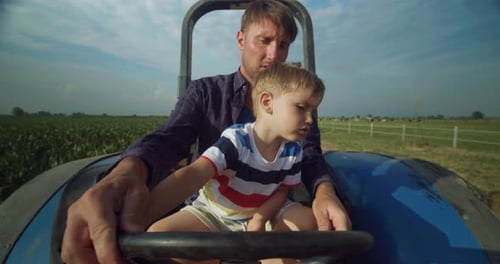 Portrait of Young Man and His Toddler Son Driving Tractor on Warm Summer Day in a Corn Field. Littl