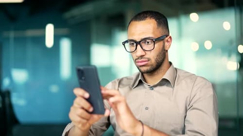 Serious focused businessman employee in glasses using a mobile phone sit at desk in modern office.