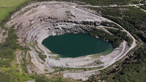 Aerial rising shot over a flooded slate quarry, showcasing steep terraced stone walls and calm green