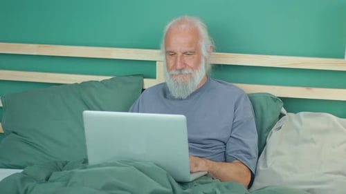 Happy Elderly Man Freelancer Works Remotely Browses the Internet Using a Laptop