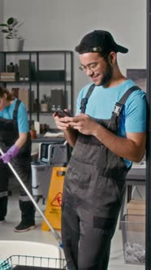 Male Cleaner Checking Smartphone while Woman Mopping Floor at Office Background