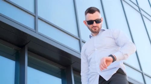 A Man Wearing Dark Glasses and a White Blue Shirt Looks at the Clock While Standing in Front of a