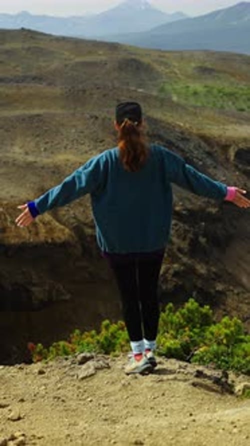 Young Woman in Casual Outdoor Clothing Stands on Rocky Mountain Edge Raising Arms in Celebration