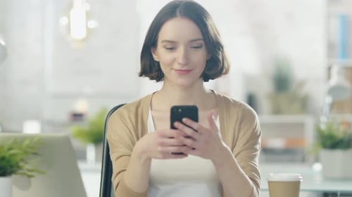 Charming Young Creative Woman Sits at Her Desk in a Bright Office and Uses Smartphone.