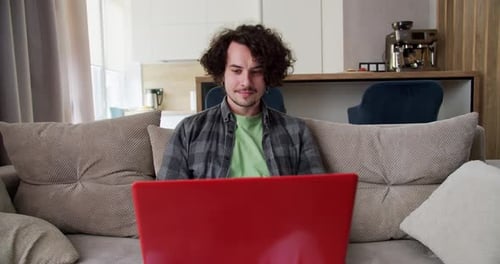 Focused Happy Brunette Guy with Curly Hair in a Gray Checkered Shirt Sits on the Sofa and Works at