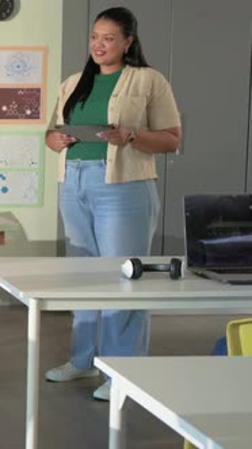 Vertical video: Camera zooming, woman in green top teaching while holding tablet in lab, PC charts
