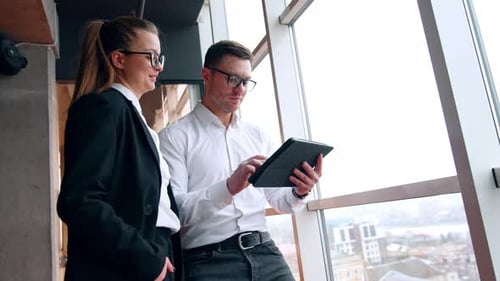 Female and male employees are in the office. Woman looks at I-pad in hands of man working on it.