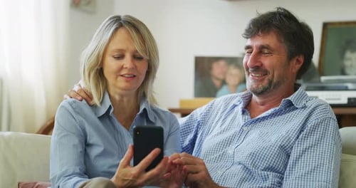A happy mature couple is using a smartphone for family entertainment while sitting on a sofa in li