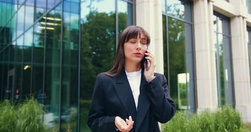 Young brunette Caucasian businesswoman in black blazer standing in front of a modern glass office bu