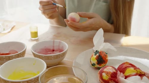 Woman decorates a marbled Easter egg with a brush, surrounded by bowls of dye and several wrapped