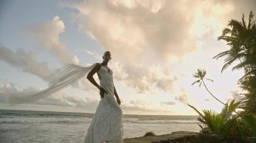 Nonbinary Black Model in Wedding Dress Poses on Tropical Beach Near Ocean at Sunset Androgynous