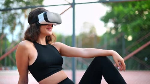Smiling woman using VR headset in a tracksuit while sitting on yoga mat on a sports field in a park