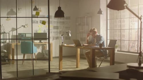 Professional Creative Designer Works on a Laptop in Loft Office, Looks at the Product Sketches and