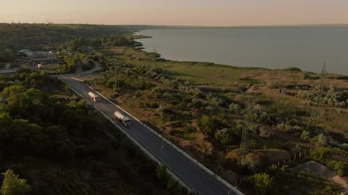 Aerial shot flying towards two white trailer trucks driving down the road, big lake in the