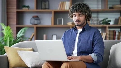 Young man typing on laptop sitting on sofa in living room at home. Freelancer works on computer