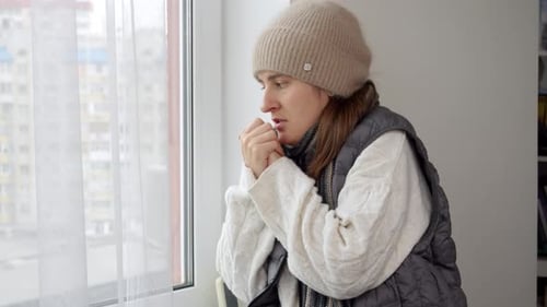 Young woman in hat and sweater freezing at home looking out of the window