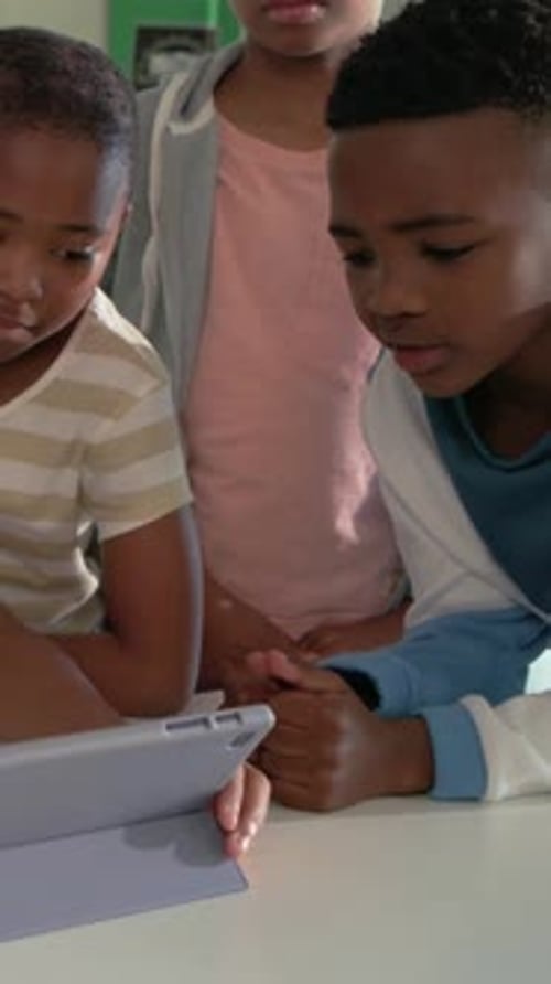 Vertical video: Reaching seated child tapping tablet on stand at school, hoodie watching to learn