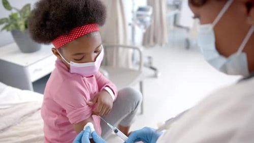 African american female doctor with face mask making injection to girl in hospital room, slow motion