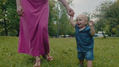 Mother Helps Her Little Son Child Take the First Step in the Park on the Grass